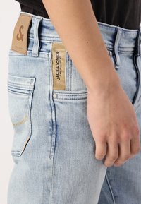 Jeans de denim azul claro con un ajuste regular, que presentan una etiqueta de cuero, costuras doradas y bolsillos traseros. La textura es suave y ligeramente desgastada.