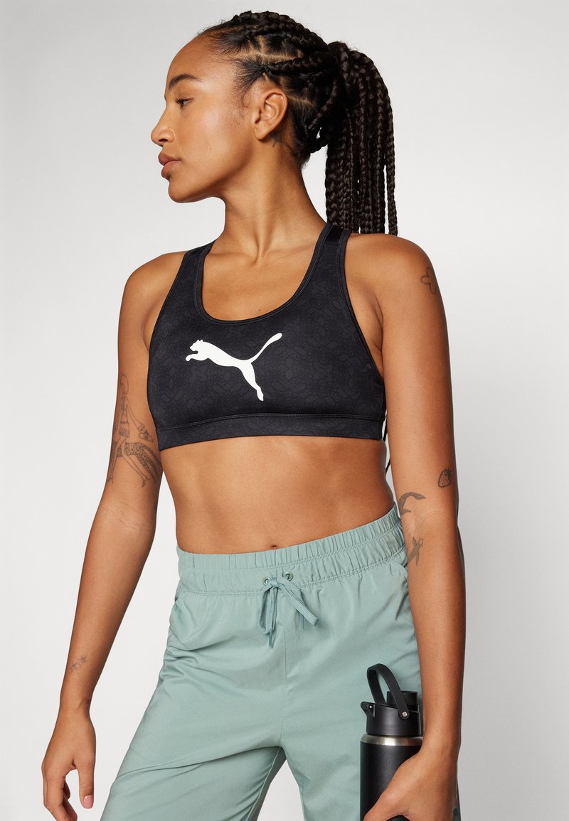 Reggiseno sportivo nero con logo Puma bianco, caratterizzato da un motivo testurizzato. Abbinato a pantaloni con cordino verde chiaro, mentre tiene in mano una borraccia nera.