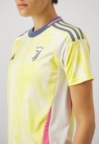 Gul mönstrad sporttröja med grå accenter, med rund halskrage, korta ärmar och ett rosa rand längs sidan. Tre stjärnor som dekoration.