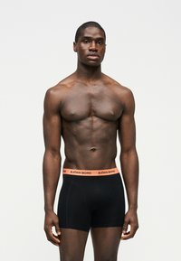 Μυώδης μαύρος άνδρας στέκεται, φορώντας μαύρο ανδρικό εσώρουχο τύπου μποξεράκι με πορτοκαλί μέση, στο οποίο αναγράφεται "Björn Borg", πάνω σε απλό ανοιχτό φόντο.