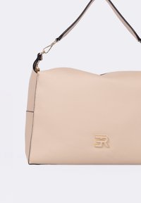 Borsa in pelle beige con superficie testurizzata, caratterizzata da una forma curva, dettagli neri e un logo dorato sul davanti. Include tracolla regolabile.