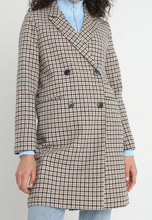 Femme portant un manteau beige à carreaux noirs à double boutonnage sur un col roulé bleu clair avec un jean, debout devant un fond uni.