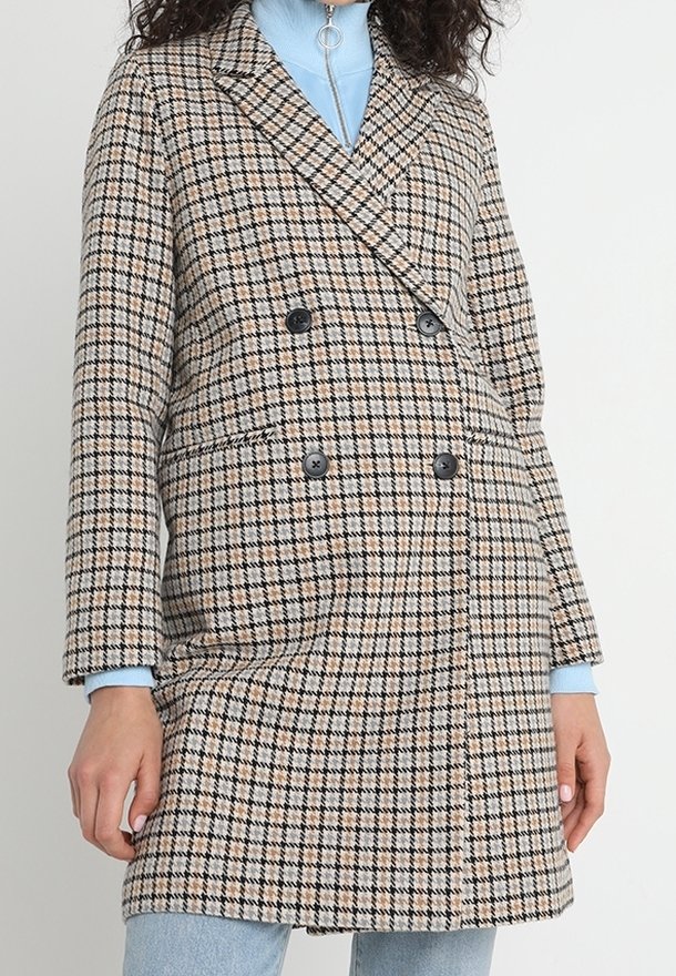 Femme portant un manteau beige à carreaux noirs à double boutonnage sur un col roulé bleu clair avec un jean, debout devant un fond uni.