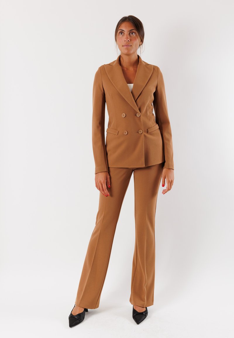 Suit marrone doppiopetto con giacca su misura e pantaloni dritti. Tessuto liscio, revers a lancia e tacchi neri eleganti.