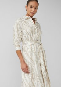 Frau trägt ein beiges und weißes gemustertes Hemdkleid mit Gürtel, dreiviertelärmeln und Knopfleiste, stehend vor einem schlichten Hintergrund.