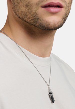 Primo piano del volto inferiore e del collo di un uomo che indossa una camicia bianca con una collana a catena d'argento con un ciondolo in cristallo nero avvolto da un serpente d'argento.