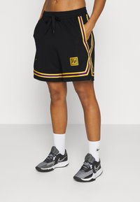 Czarne sportowe szorty z fioletowo-złotymi paskami, wyposażone w boczne kieszenie i złotą naszywkę z logo. Do tego szare buty Nike i białe skarpetki.