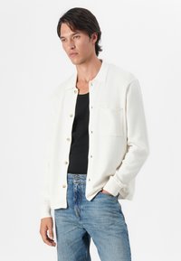 Witte gebreide cardigan met een kraag, met gouden knopen en een voorzak. Gepaard met een zwarte tanktop en blauwe denim jeans.
