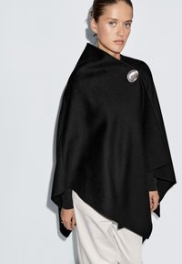 Žena v černém asymetrickém plášti zapnutém velkou stříbrnou broží, v kombinaci s bílými kalhotami, na jednoduchém světlem pozadí.