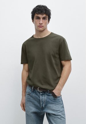 Junger Mann mit lockigem dunklem Haar, der ein olivegrünes T-Shirt und hellblaue Jeans mit einem braunen geflochtenen Gürtel trägt, vor einem einfachen Hintergrund stehend.