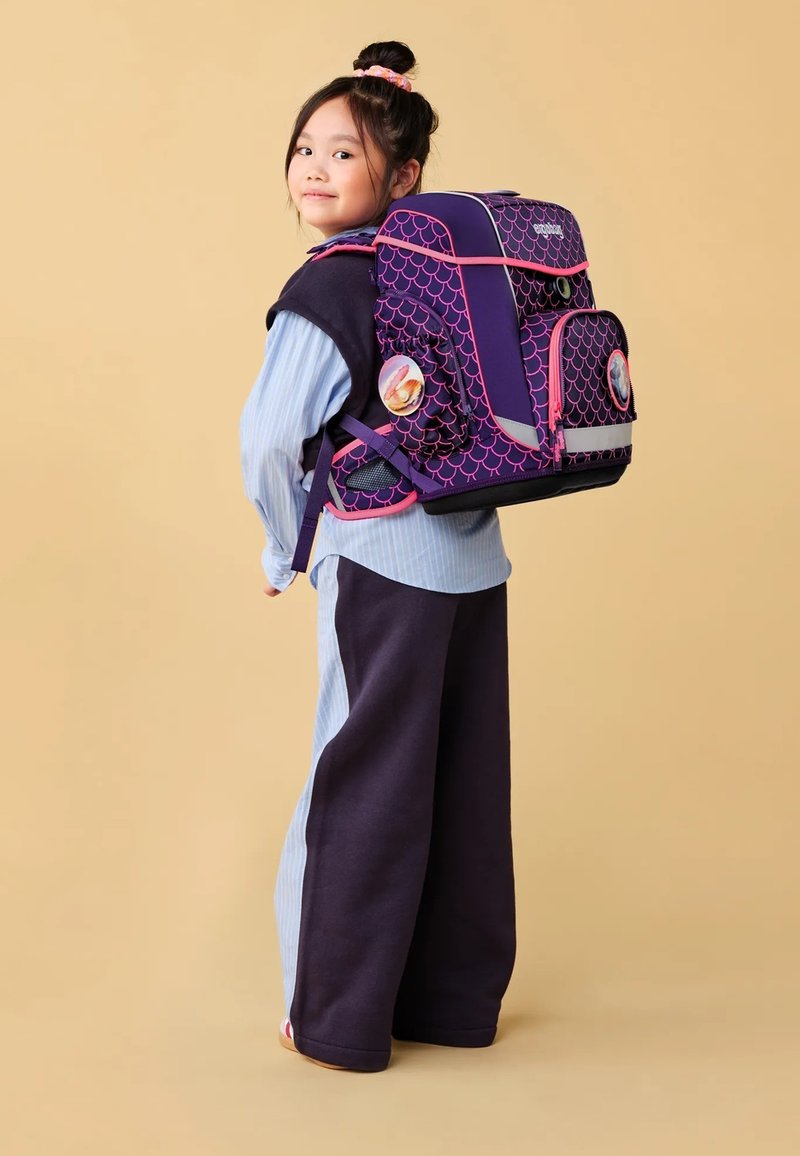 Lila- und rosa gemusterter Rucksack mit Fischschuppen-Design, mehreren Taschen und runden Abzeichen, getragen von einem Kind in einem gestreiften blauen Oberteil.