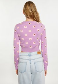 Pulover scurt liliachiu cu flori albe și centrul roșu, cu manșete și tiv ondulat. Asortat cu blugi largi de culoare albastră deschis, vizibil din spate.