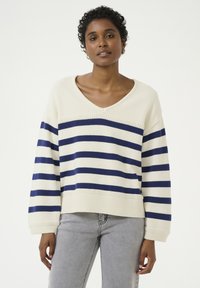 Krem pulover iz pletenine z mornarskimi črtami, z V-izrezom, rebrastimi manšetami in robom, prikazan z sivo denim hlačami. Tekstura deluje mehko in udobno.