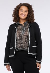 Černý cardigan se stříbrným lemem a detaily s třásněmi, vrstvený přes černobílé struktuované blůze. Obě části jsou strukturované, ale zároveň jemné.