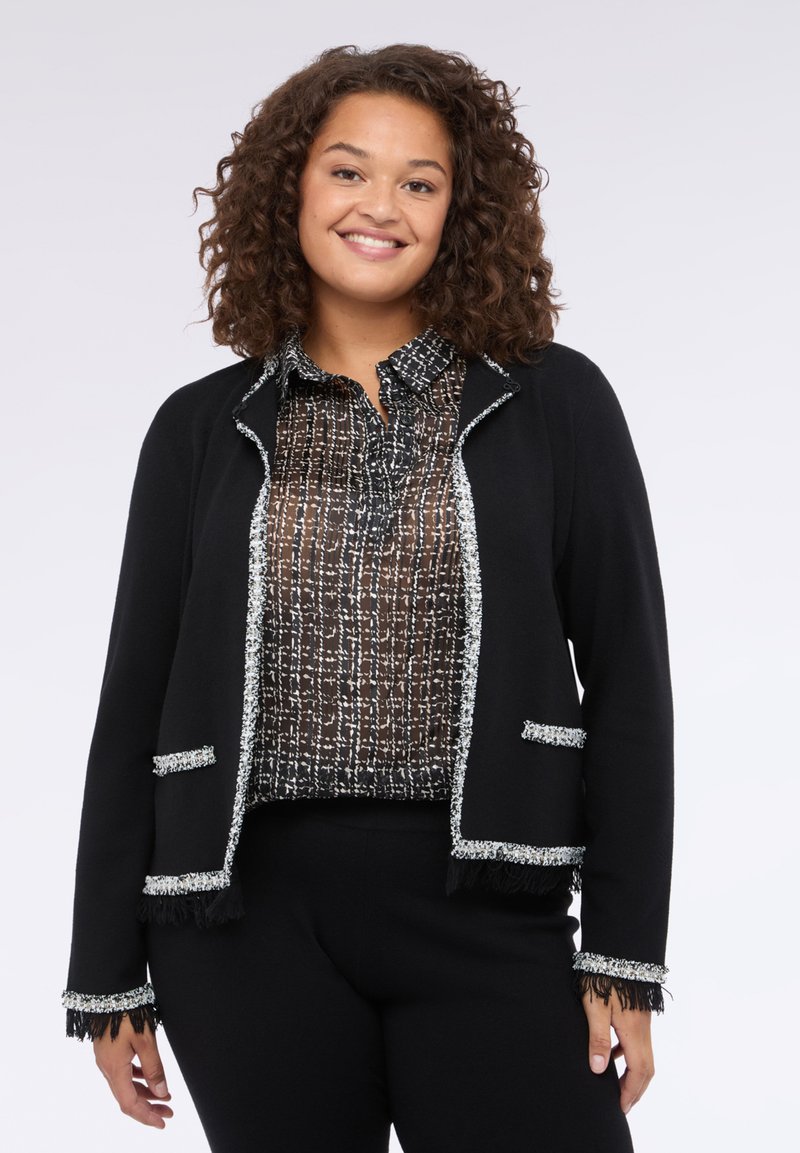 Černý cardigan se stříbrným lemem a detaily s třásněmi, vrstvený přes černobílé struktuované blůze. Obě části jsou strukturované, ale zároveň jemné.