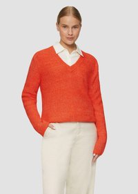 Oranger Pullover mit V-Ausschnitt, gerippter Textur und langen Ärmeln, kombiniert mit einem weißen Hemd mit Kragen und hellen Hosen.