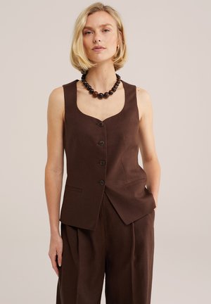 Žena oblečená v bezrukávovej hnedej vestke na gombíky a zodpovedajúcich nohaviciach s vysokým pásom, so tmavým korálkovým náhrdelníkom, stojaca pred jednoduchým pozadím.