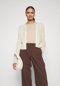 Cream knitted cardigan with a textured pattern, featuring a cropped design and open front. Paired with a beige top and brown trousers.