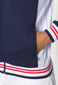 Chaqueta deportiva azul marino y blanca con cierre de cremallera, patrón de rayas finas y puños con franjas rojas. Cuenta con bolsillos laterales y herrajes de marca.