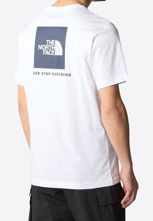 Mann trägt weißes T-Shirt mit großem dunkelblauem Quadrat und "THE NORTH FACE"-Logo sowie unvollständigem Text "NEVER STOP EXPLORING" auf der Rückseite.