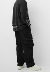 Černé oversized tričko přehnuté přes černou dlouhoupážou, v kombinaci s volnými černými cargo kalhotami s několika kapsami. Bílé tenisky.