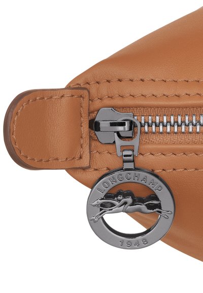 Ecke einer beigefarbenen Ledertasche mit silbernem Reißverschluss und markantem Anhänger mit springendem Hundemotiv. Nahtdetails entlang der Kante.