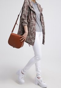 Kamouflage-mönstrad jacka, grå t-shirt, vita skinny jeans, brun crossbody-väska och vita platåsneakers.