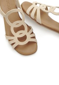 Beige suede sandals featuring circular cutouts, multiple straps, and a flat brown sole with a smooth texture.