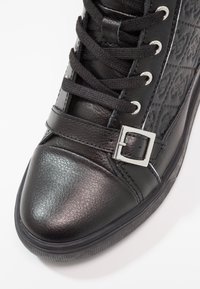Black leather sneaker with textured side panel, round toe, flat sole, and fabric laces. Includes a silver buckle accent on the strap.