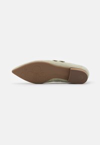 Chaussure en suède beige avec un bout pointu et une semelle en caoutchouc brun clair. Présente du matériel décoratif sur le côté et un logo en relief sur la semelle.
