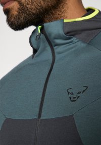 Férfi cipzáras dzseki zöld és fekete színben, magas gallérral, diszkrét logóval és kontrasztos neon sárga részletekkel a gallér szegélyén.