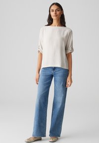 Beige Leinenoberteil mit Bootsausschnitt und hochgerollten Ärmeln, kombiniert mit hellblauen, weit geschnittenen Jeans und beige flachen Schuhen. Insgesamt lässiges Outfit.