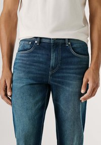 Vaqueros de mezclilla azul con corte recto, con cremallera, trabillas y bolsillos delanteros. Desvanecido ligero y detalles de costuras blancas.