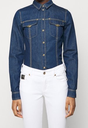 Osoba nosząca dopasowaną, ciemnoniebieską jeansową koszulę z złotymi guzikami oraz białe spodnie z złotym guzikiem i małą czarną metką.