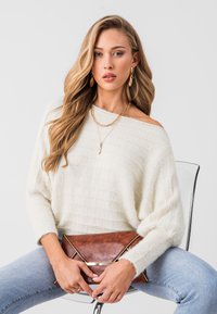 Suéter de punto en crema claro con textura acanalada, combinado con jeans azules. Sostiene un bolso de mano de cuero marrón con acentos dorados.