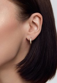 Złoty kolczyk w kształcie obręczy o błyszczącej powierzchni, z małymi, przezroczystymi kamieniami osadzonymi po jednej stronie. Noszony na uchu w zbliżeniu, podkreślającym wzór.