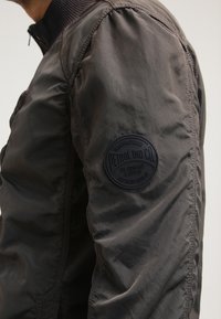 Grå nylonjakke med ribbet krave, der har en rund sortapel på ærmet, hvor der står "Petrol Ind Co." Struktureret overflade med let glans.