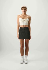 Kreeme kollane heegeldatud halter top, kombineerituna tume rohelise mini seeliku ja eest taskutega. Mudel kannab valgeid tossusid ja sokke.