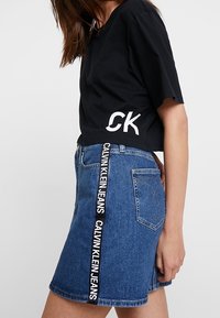 Kvinna klädd i svart kortärmad t-shirt med vitt "CK"-logotyp och blå denimkjol med vertikal Calvin Klein Jeans-branding på sidan.