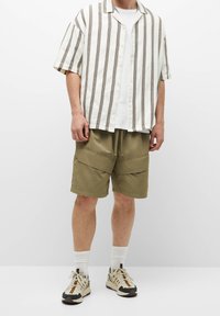 Camisa blanca de manga corta con rayas verticales marrones, combinada con pantalones cortos cargo verdes oliva con grandes bolsillos y cintura con cordón.