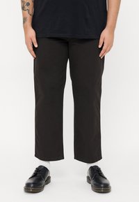 Black, cropped trousers made of durable fabric, featuring a straight cut and minimal stitching. Paired with dark shoes and white socks.