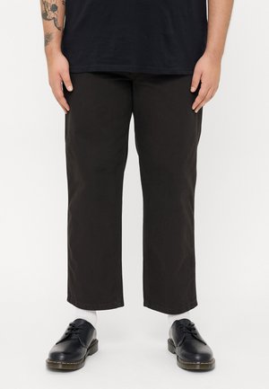 Zwarte, cropped broeken van duurzaam materiaal, met een rechte snit en minimale stiksels. Gecombineerd met donkere schoenen en witte sokken.