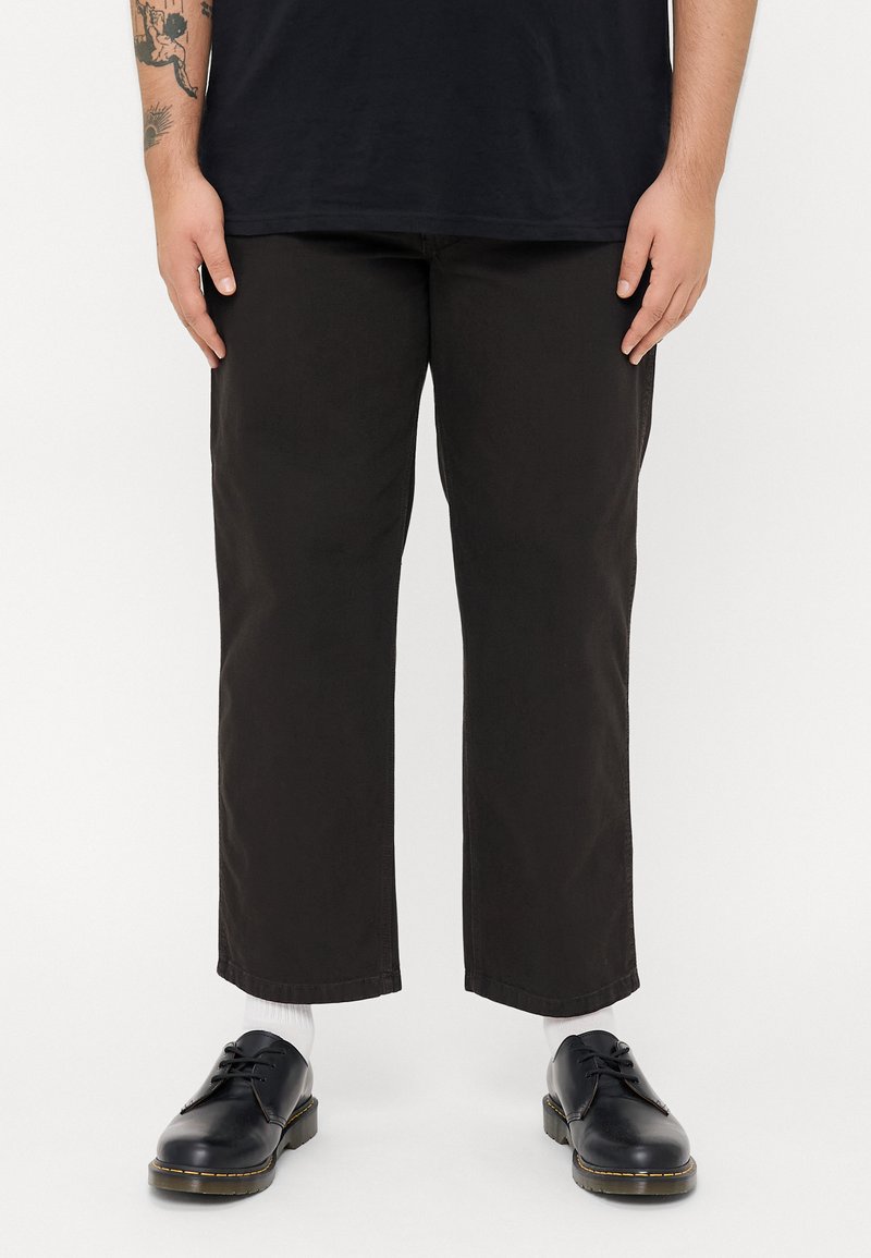Zwarte, cropped broeken van duurzaam materiaal, met een rechte snit en minimale stiksels. Gecombineerd met donkere schoenen en witte sokken.