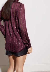 Blusa a maniche lunghe bordeaux in tessuto trasparente, con pois dorati e righe verticali. Presenta un yoke sul retro e polsini con bottoni.
