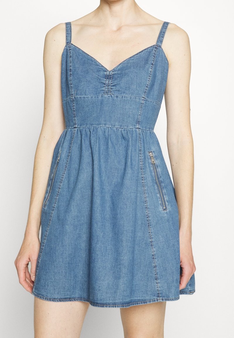 Femme portant une robe en denim bleu sans manches avec un décolleté en cœur, un corsage ajusté, une jupe froncée et des poches latérales zippées.