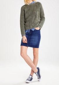 Olivgrön stickad tröja med strukturerade mönster, över en ljusblå skjorta med krage. Mörk denim blyertskjol och marinblå sneakers.