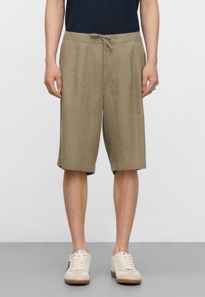 Mężczyzna w beżowych krótkich spodenkach do kolan ze sznurkiem w pasie, czarnej koszulce i kremowych trampkach, stojący na szarej podłodze na jednolitym tle.