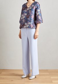 Floral gemusterte, glänzende Bluse in lila-blauen Tönen mit kurzen Puffärmeln. Kombiniert mit hell-lila weit geschnittenen Hosen und silbernen spitzen High Heels.