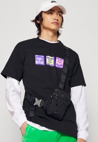 Svart t-shirt med tre färgglada grafiska designer, ihop med en svart midjeväska och ljusgröna byxor. Vit långärmad undertröja och keps.