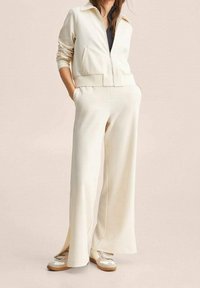 Giacca zip con collo di colore crema, abbinata a pantaloni larghi di crema. Caratterizzata da un tessuto morbido e tasche laterali; le scarpe sono con accenti bianchi e beige.